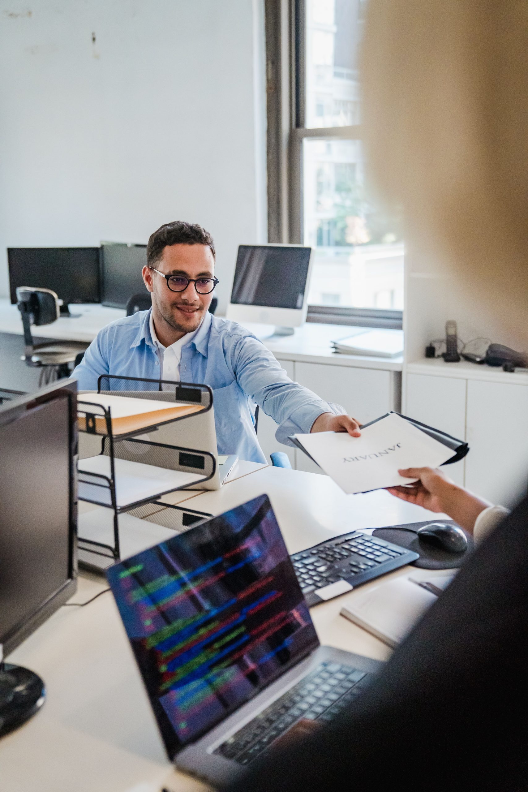 Two office workers exchanging documents in a bright office setting.